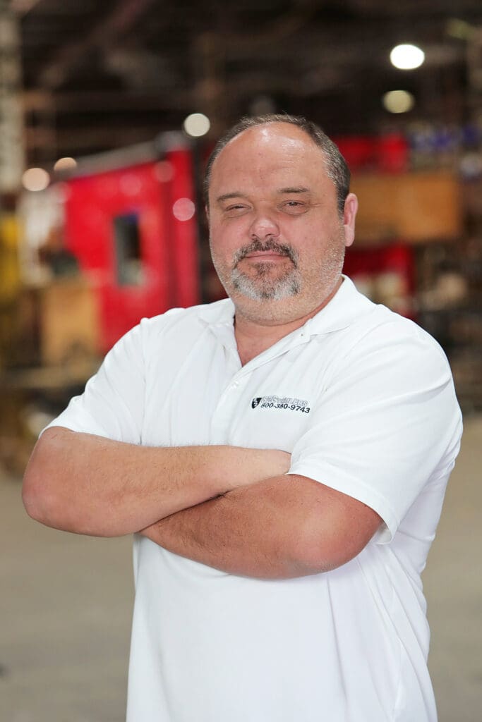 A man with a beard and mustache, wearing a white polo shirt with his arms crossed, standing indoors with a blurred industrial-looking background.
