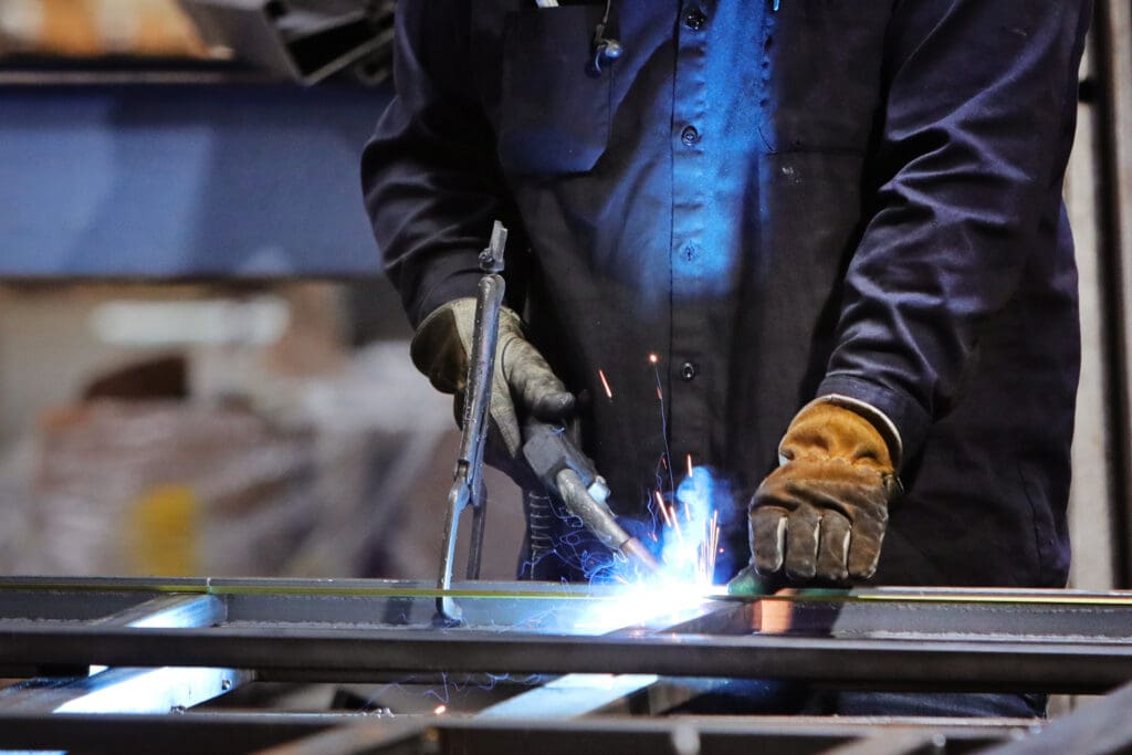 A close-up shot of a person wearing protective gloves and dark clothing, performing welding on a metal surface, with bright blue sparks and light emanating from the welding point.